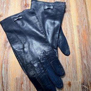 May include: A pair of dark navy blue leather gloves featuring a smooth finish and a small, centered decorative bow detail on the cuff of each glove. The gloves are constructed with visible stitching along the fingers and a classic wrist-length cut.