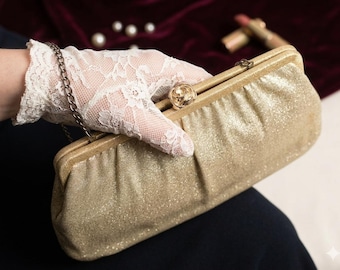 May include: A gold glittery clutch bag featuring a metallic frame and a rose-shaped clasp. The rectangular evening bag includes a thin silver-tone chain strap and is held by a hand wearing a white lace glove with a floral pattern.