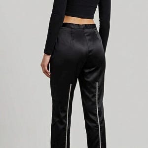 Puede incluir: Un top negro de manga larga y pantalones de sat&eacute;n negro con rayas verticales blancas. Los pantalones son tobilleros y se combinan con botas de plataforma negras. El conjunto es elegante y moderno.