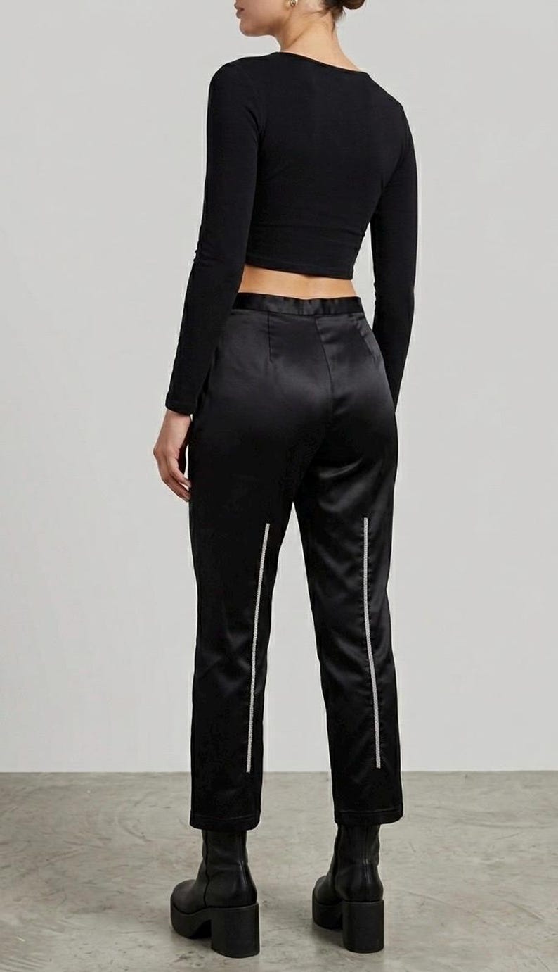 Puede incluir: Un top negro de manga larga y pantalones de sat&eacute;n negro con rayas verticales blancas. Los pantalones son tobilleros y se combinan con botas de plataforma negras. El conjunto es elegante y moderno.