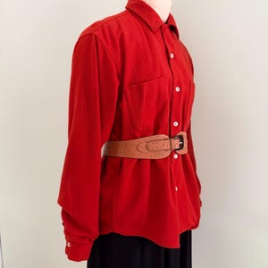 May include: A long-sleeved, solid red button-down shirt featuring a pointed collar and two chest pockets, cinched at the waist with a wide, tan-colored textured belt that has a metal buckle and a distinct dotted pattern across its surface.