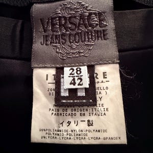 Puede incluir: Primer plano de una etiqueta negra de Versace Jeans Couture. La etiqueta es gris con el nombre y el logotipo de la marca. Incluye informaci&oacute;n sobre la talla, la composici&oacute;n de la tela y el texto "Hecho en Italia."