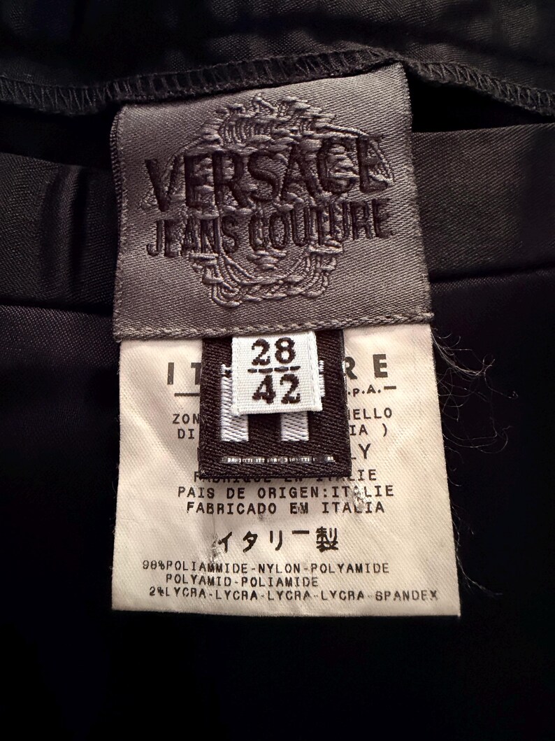 Puede incluir: Primer plano de una etiqueta negra de Versace Jeans Couture. La etiqueta es gris con el nombre y el logotipo de la marca. Incluye informaci&oacute;n sobre la talla, la composici&oacute;n de la tela y el texto "Hecho en Italia."