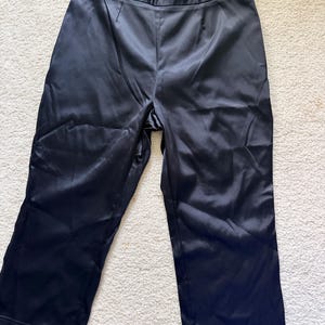Puede incluir: Pantalones de sat&eacute;n negro con corte de pierna ancha. Los pantalones tienen cintura alta y cierre de bot&oacute;n. La tela tiene un ligero brillo, reflejando la luz. Los pantalones est&aacute;n colocados sobre una superficie blanca.