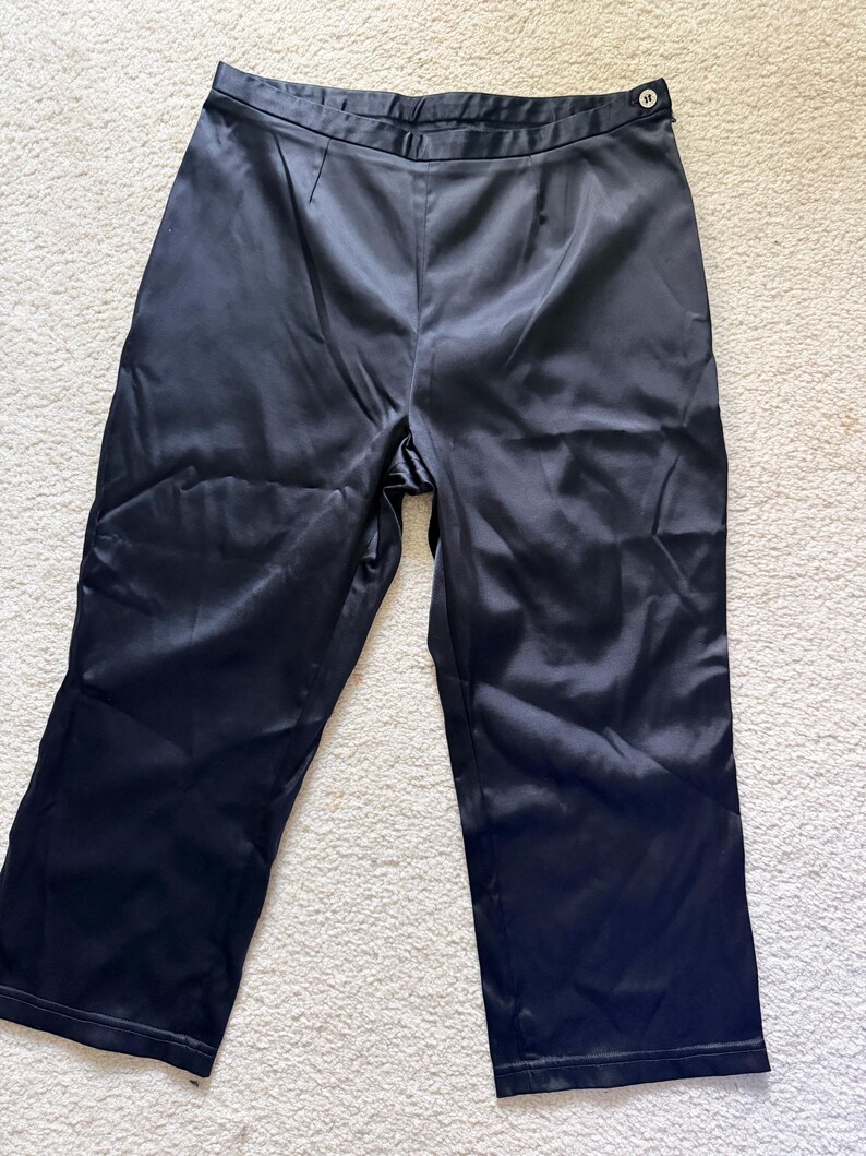 Puede incluir: Pantalones de sat&eacute;n negro con corte de pierna ancha. Los pantalones tienen cintura alta y cierre de bot&oacute;n. La tela tiene un ligero brillo, reflejando la luz. Los pantalones est&aacute;n colocados sobre una superficie blanca.