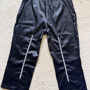 Puede incluir: Pantalones de sat&eacute;n negro con cintura alta y pernera recta. Los pantalones tienen una raya vertical blanca en la parte delantera de cada pernera. La tela tiene un ligero brillo.