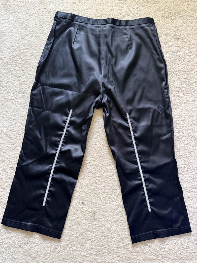 Puede incluir: Pantalones de sat&eacute;n negro con cintura alta y pernera recta. Los pantalones tienen una raya vertical blanca en la parte delantera de cada pernera. La tela tiene un ligero brillo.
