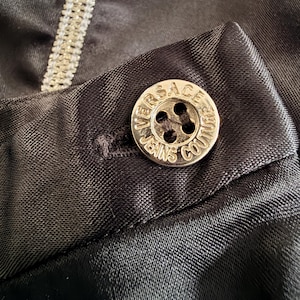 Puede incluir: Primer plano de una tela de sat&eacute;n negro con un bot&oacute;n plateado. El bot&oacute;n redondo lleva grabada la inscripci&oacute;n "VERSACE JEANS COUTURE". La tela tiene un brillo sutil.