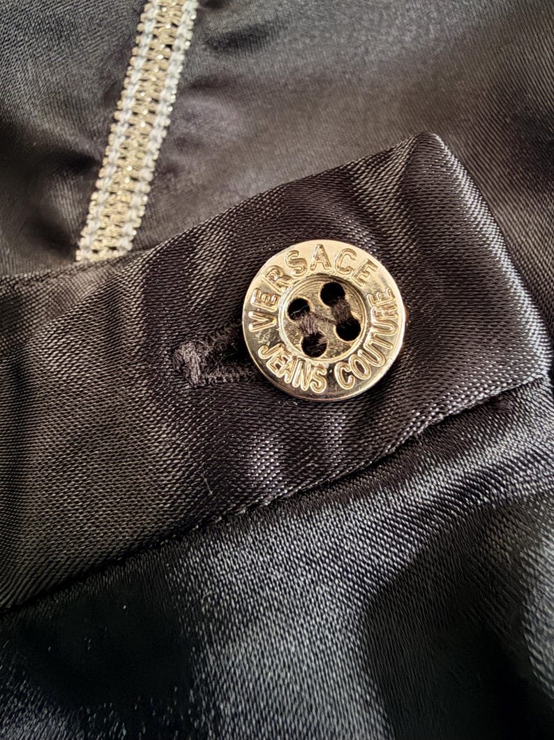 Puede incluir: Primer plano de una tela de sat&eacute;n negro con un bot&oacute;n plateado. El bot&oacute;n redondo lleva grabada la inscripci&oacute;n "VERSACE JEANS COUTURE". La tela tiene un brillo sutil.
