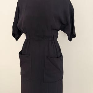 May include: A solid black dress featuring a round neckline, short sleeves, and an elasticized waistline. The garment includes two large, square patch pockets positioned on the front of the skirt section, providing a functional and structured appearance.