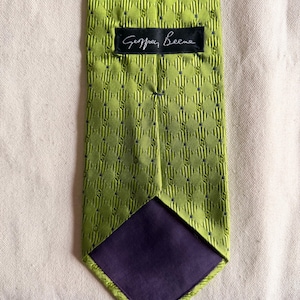 May include: A lime green necktie featuring a repeating diamond pattern with small dark blue dots and a textured ribbed finish. A black fabric label with the white text Geoffrey Beene is stitched near the top, and the bottom lining is dark purple.