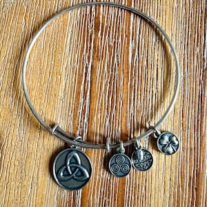May include: A silver-toned metal bangle bracelet featuring four dangling charms. The charms include a circular triquetra symbol, a small triskelion, a rectangular tag with the text IRELAND, and a four-leaf clover shape with a dark oxidized finish.