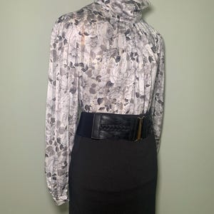 May include: A floral print blouse with a high neck and long sleeves, paired with a black skirt and a wide black belt with a gold-tone buckle. The blouse features a pattern of gray roses on a white background.