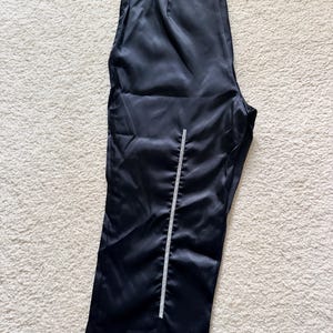 Puede incluir: Pantalones de sat&eacute;n negro con un detalle vertical de pedrer&iacute;a en la parte delantera. Los pantalones tienen una cintura alta y una pernera ancha. La tela tiene un brillo sutil y est&aacute;n sobre una alfombra clara.