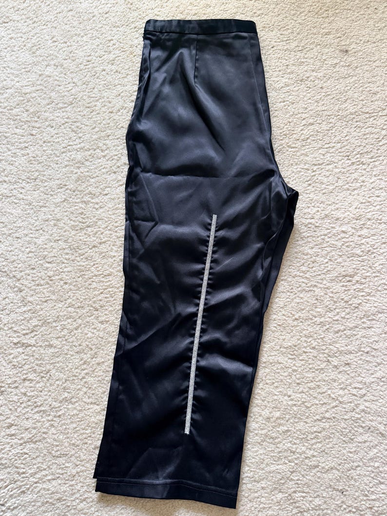 Puede incluir: Pantalones de sat&eacute;n negro con un detalle vertical de pedrer&iacute;a en la parte delantera. Los pantalones tienen una cintura alta y una pernera ancha. La tela tiene un brillo sutil y est&aacute;n sobre una alfombra clara.