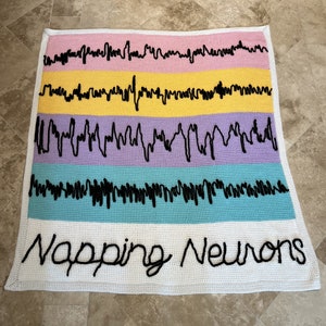 Puede incluir: Una manta de crochet blanca con un patrón de rayas de colores y el texto "Napping Neurons" en hilo negro.