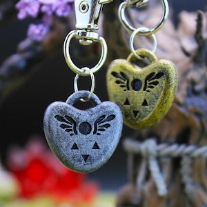 May include: Two heart-shaped keyrings with a wing and triangle design. One is silver, the other gold. Both are attached to silver key rings and a silver clasp.