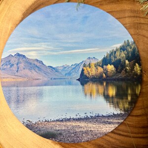 May include: A scenic landscape photograph featuring a lake with mountains in the background. The lake is calm and reflects the sky and mountains. The mountains are covered in trees and snow. The sky is blue with white clouds.