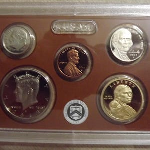 May include: A set of United States coins in a clear plastic case. The coins include a dime, a penny, a half dollar, and two other coins. The coins are set against a brown background.