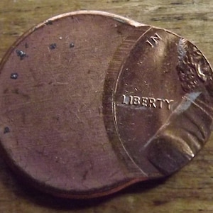 May include: A copper-colored US penny coin with a damaged edge. The coin is showing the obverse side with the words "LIBERTY" and "IN GOD WE TRUST" visible. The coin is on a wooden surface.