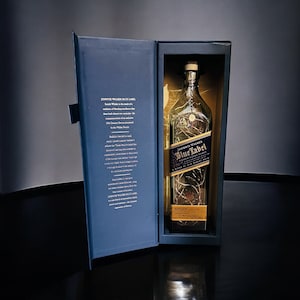 Op de afbeelding: Een blauwe en gouden doos met een heldere glazen fles Johnnie Walker Blue Label Scotch Whisky. De fles is versierd met een gouden etiket en een gouden dop. De doos heeft een wit etiket met tekst over de whisky.
