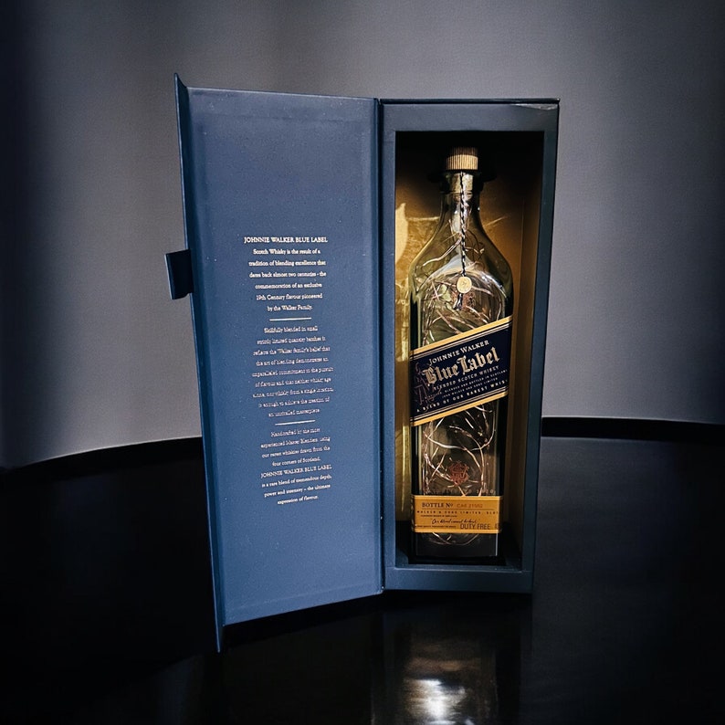 Op de afbeelding: Een blauwe en gouden doos met een heldere glazen fles Johnnie Walker Blue Label Scotch Whisky. De fles is versierd met een gouden etiket en een gouden dop. De doos heeft een wit etiket met tekst over de whisky.