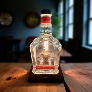 May include: A repurposed Jack Daniel's whiskey bottle transformed into a lamp. The clear glass bottle features the brand's logo and text, with a red label. The lamp sits on a dark wooden base and has a visible filament bulb.