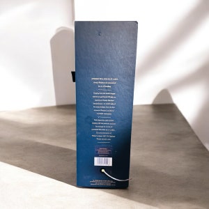 Op de afbeelding: Een blauwe kartonnen doos met gouden letters die "JOHNNIE WALKER BLUE LABEL" lezen en de Schotse whisky beschrijven als de top van de kunst van het blenden. De doos vermeldt ook dat alleen de beste vaten voor deze whisky worden geselecteerd.