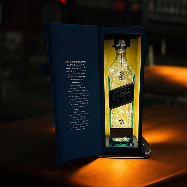 Op de afbeelding: Een doorzichtige glazen fles Johnnie Walker Blue Label Scotch whisky met een lichtsnoer erin. De fles zit in een houten doos met een label waarop "Johnnie Walker Blue Label" staat.