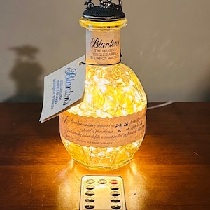 May include: A clear glass bottle with a label that reads "Blanton's The Original Single Barrel Bourbon Whiskey". The bottle is decorated with a horse and jockey figurine on top and has a string of lights inside. The bottle is sitting on a wooden surface with a remote control for the lights.
