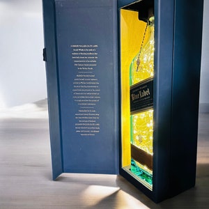 Op de afbeelding: Een blauwe en gouden geschenkdoos met een fles John Walker & Sons Blue Label Blended Scotch Whisky. De fles is verlicht met witte kerstverlichting.