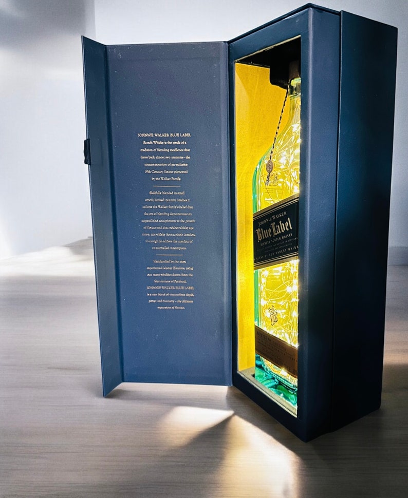 Op de afbeelding: Een blauwe en gouden geschenkdoos met een fles John Walker & Sons Blue Label Blended Scotch Whisky. De fles is verlicht met witte kerstverlichting.