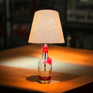 May include: A table lamp made from a clear glass bottle with a tan fabric lampshade. The bottle has a red cap and a label that reads "The Quarter Barrel Whiskey".