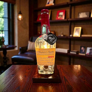 May include: A clear glass Maker's Mark bottle with a red wax seal and a black tag. The label is orange and features text including "Wood Finishing Series" and "2024". The bottle is displayed on a dark wooden stand.