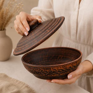 May include: A dark brown clay bowl with a matching lid, featuring a textured exterior with a repeating leaf-like pattern around the base. The bowl has a wide, circular opening and a smooth interior, while the lid has a central knob handle.