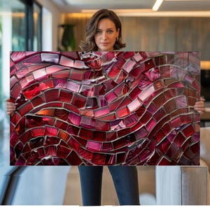 May include: A rectangular mosaic artwork featuring a flowing design in shades of red, burgundy, and pink. The mosaic is composed of small, irregularly shaped tiles, creating a textured and dynamic visual effect. The artwork is held up in front of a person.