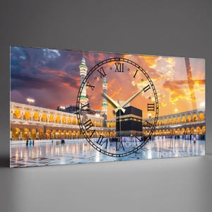Può includere: Un orologio da parete rettangolare con un'immagine vibrante della Masjid al-Haram alla Mecca, in Arabia Saudita, con un quadrante sovrapposto. Il quadrante ha numeri romani e lancette dorate, su uno sfondo di cielo al tramonto.