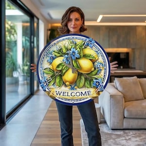 Op de afbeelding: Ronde wandklok met een blauw-witte rand, met een citroen- en bloemmotief. De wijzerplaat toont Romeinse cijfers en het woord "WELCOME" op een banner. Een decoratief woonaccessoire.