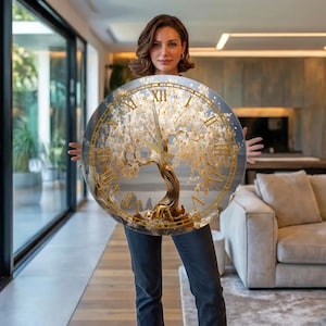 May include: A circular wall clock featuring a gold-toned tree with white blossoms as the central design. The clock face displays gold Roman numerals around the perimeter and includes gold-colored hour and minute hands centered on the tree trunk.
