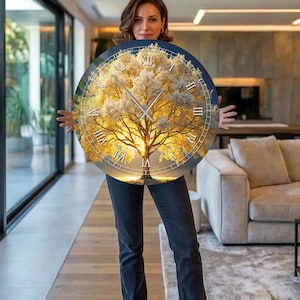 Op de afbeelding: Een ronde wandklok met een boomontwerp en een gouden zonsondergang achtergrond. De klok heeft Romeinse cijfers en zilveren wijzers. De klok wordt vastgehouden door een persoon in een moderne woonkamer.