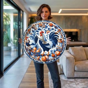 May include: A round decorative plate with a rooster and hen design in blue and white. The plate features a detailed illustration of a rooster, hen, eggs, and floral elements. The plate has a blue border with orange accents.