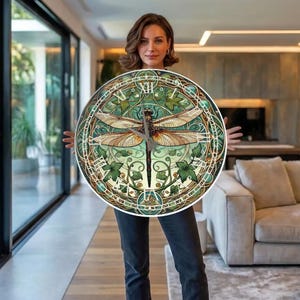 Könnte beinhalten: Runde Wanduhr mit Libellen-Design. Das Zifferblatt hat ein grünes und beiges Farbschema mit einer Libelle in der Mitte und dekorativen Efeublättern am Rand. Die Zeiger der Uhr sind schwarz.