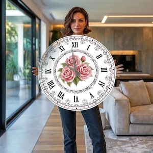 May include: A round wall clock featuring a white face with black Roman numerals and a decorative black filigree border. The centre of the clock displays a cluster of three pink roses with green leaves and two grey clock hands.