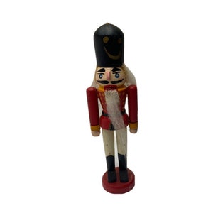 May include: A wooden nutcracker figurine with a red and gold uniform, a black hat with a smiley face, and a white beard.