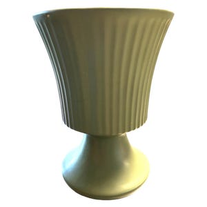 May include: A sage green, pedestal vase with a fluted, urn-shaped top. The vase has a rounded base and a smooth, matte finish. The overall design is simple and elegant.