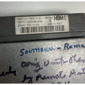 May include: A gray electronic control module with a white label featuring text and a barcode. The label includes the text "EEC-V MPC-176" and "HBM1". Handwritten text in blue ink is visible on the metal casing.