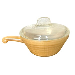 May include: A vintage, amber-colored Pyrex casserole dish with a clear glass lid. The dish has a handle and a ribbed design. The lid has a small, textured handle. The dish is designed for cooking and serving.