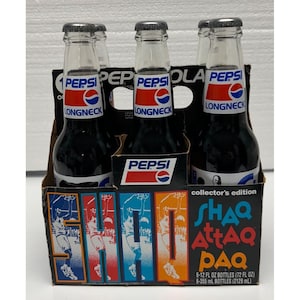 May include: A six-pack of Pepsi Longneck bottles in a cardboard carrier. The carrier features a colorful design with basketball imagery and the text "SHAQ Attaq PAQ" and "collector's edition". Each bottle has a Pepsi label.