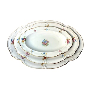 May include: A set of three white oval serving platters with a floral design. Each platter has a gold rim and a scalloped edge. The floral pattern includes pink, blue, and yellow flowers.