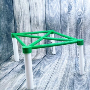 May include: A small, geometric structure with a green frame and white cylindrical legs. The frame is a series of connected triangles. The legs are evenly spaced and appear to be made of plastic. The structure is on a wooden surface.